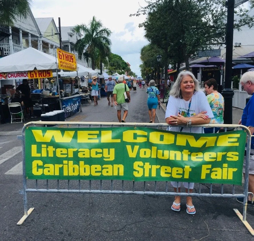 A woman standing behind a green banner that reads "Welcome Literacy Volunteers Caribbean Street Fair" on a busy street with tents, people, and trees in the background.