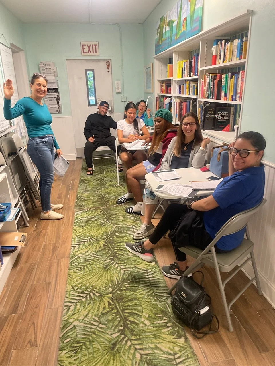 A group of seven women and one man sitting in a small room with bookshelves, a green leaf-patterned rug, and a woman standing waving and smiling.