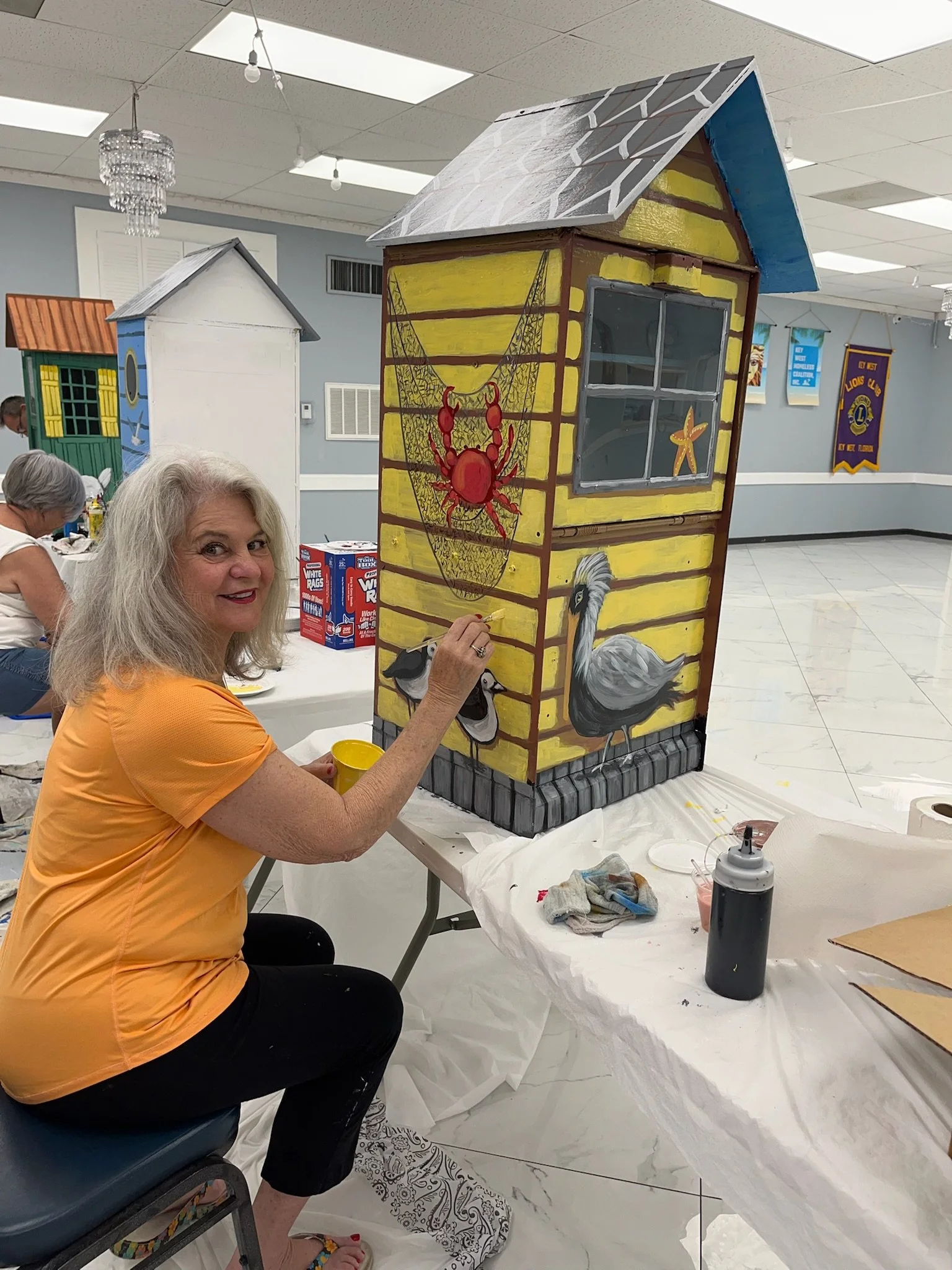 Woman painting a colorful, detailed mural on a small, house-shaped structure, featuring a crab, a starfish, a crane, and a bird, inside a community center or event hall with other art projects and banners in the background.