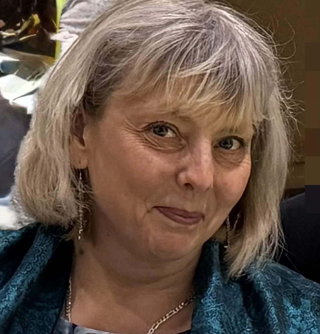 Close-up of a middle-aged woman with blonde hair, blue eyes, and a smile, wearing a teal top and jewelry.