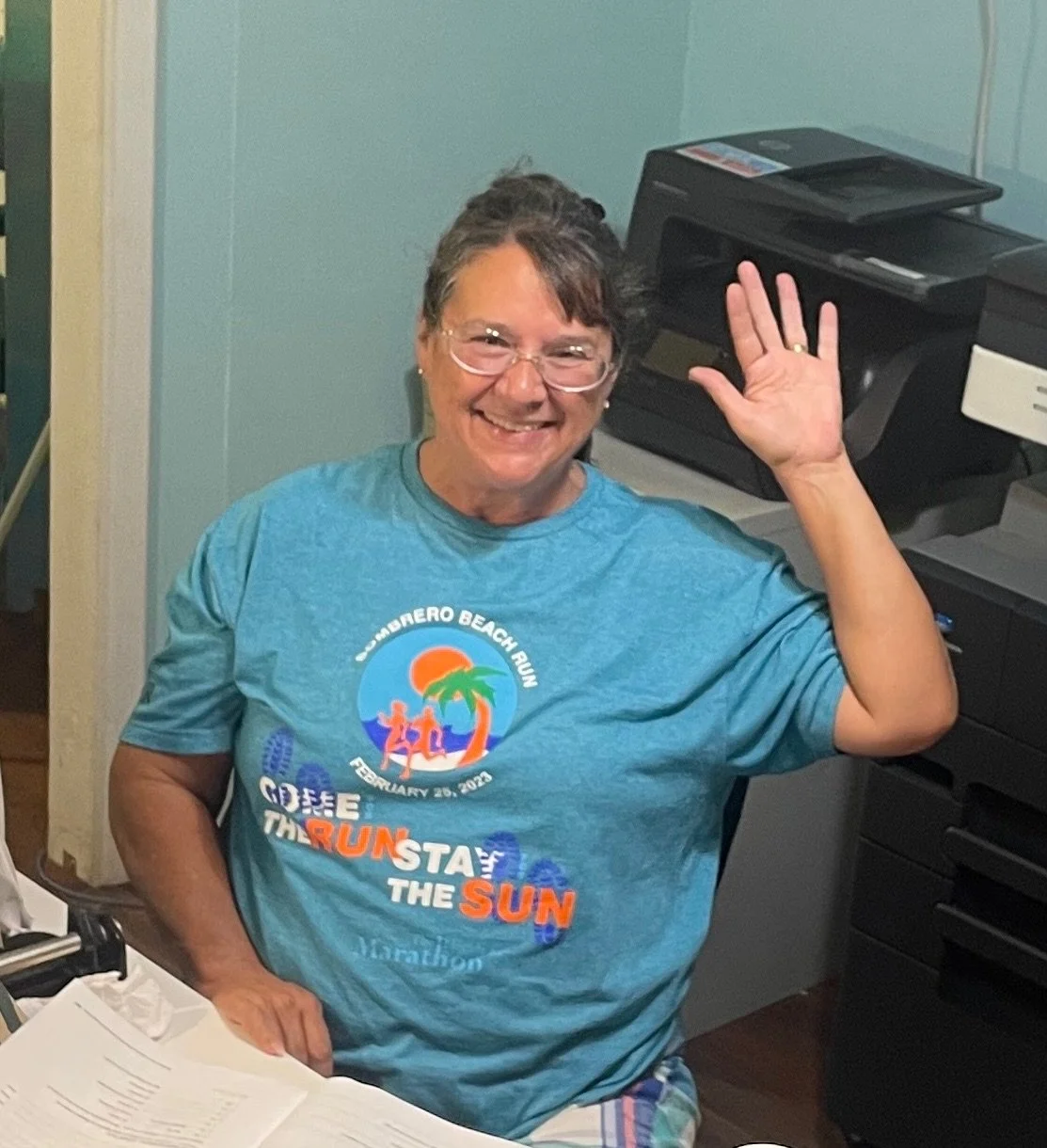 A smiling woman with glasses, wearing a blue T-shirt that says 'Run the Sun' and features a sunset scene. She is waving and sitting at a desk with papers, with a printer behind her.