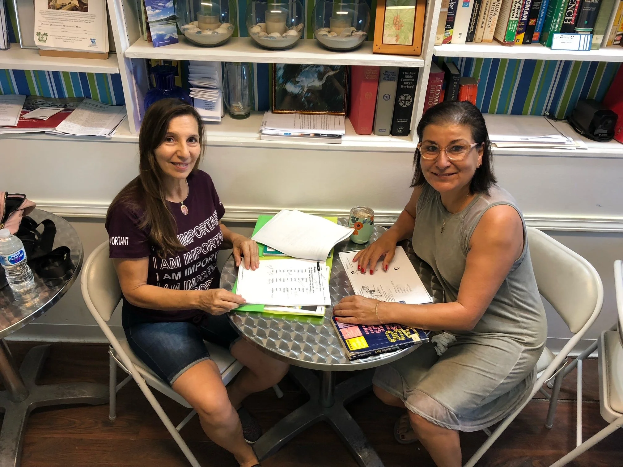Two women sitting at a table with notebooks and papers, smiling, in a room with bookshelves and decorative items behind them.
