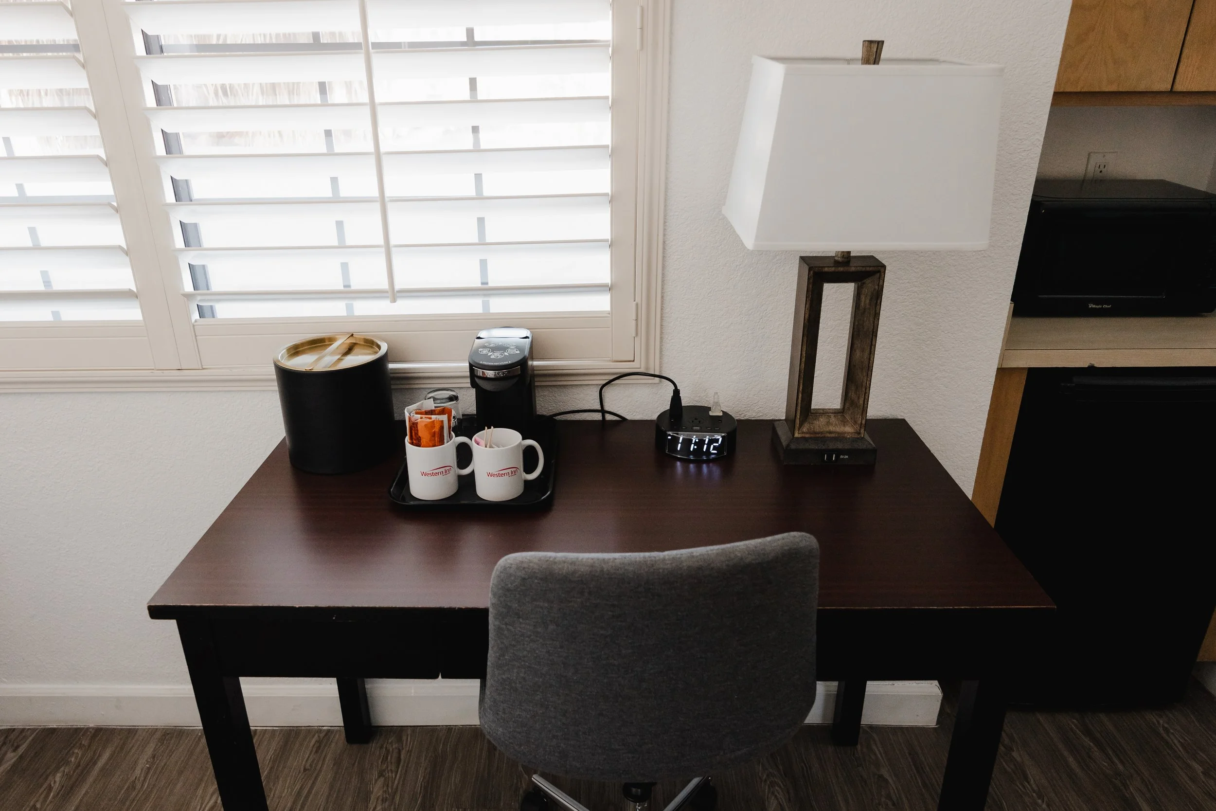 A dark brown wooden table with a gray upholstered chair in front of it, positioned near a window with white blinds. On the table, there is a black tray holding two white coffee mugs with a red logo that says 'Western Inn,' one of which contains coffee stirrers. There is also a black electric kettle, a small jar of creamer, and a black digital clock displaying '11:12'. A table lamp with a rectangular wooden base and white shade is also on the table. To the right, part of a kitchen counter with a black microwave oven and a white electrical outlet on the wall can be seen.