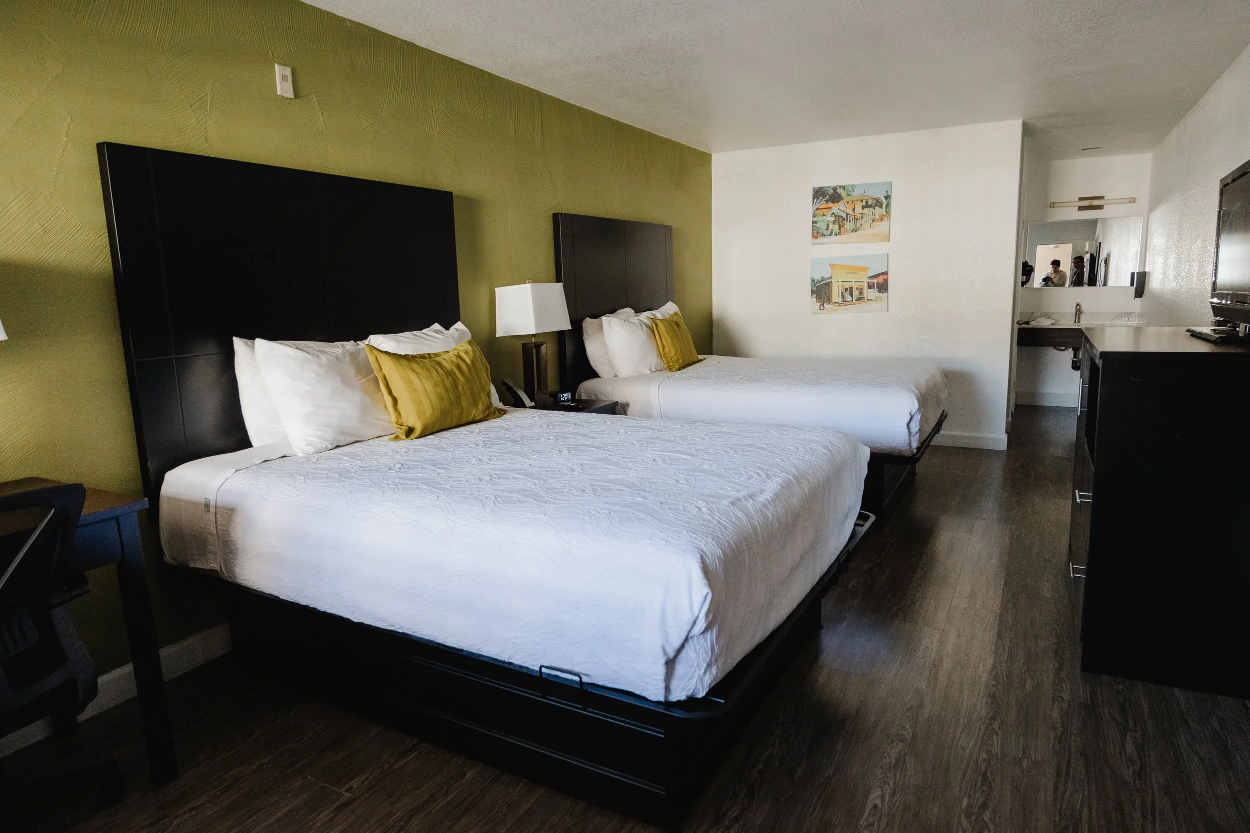 Two queen beds in a hotel room with dark headboards, white bedding, and yellow pillows, a nightstand with a lamp and clock, a wall with paintings, and a partial view of a kitchenette with a mirror reflecting two people