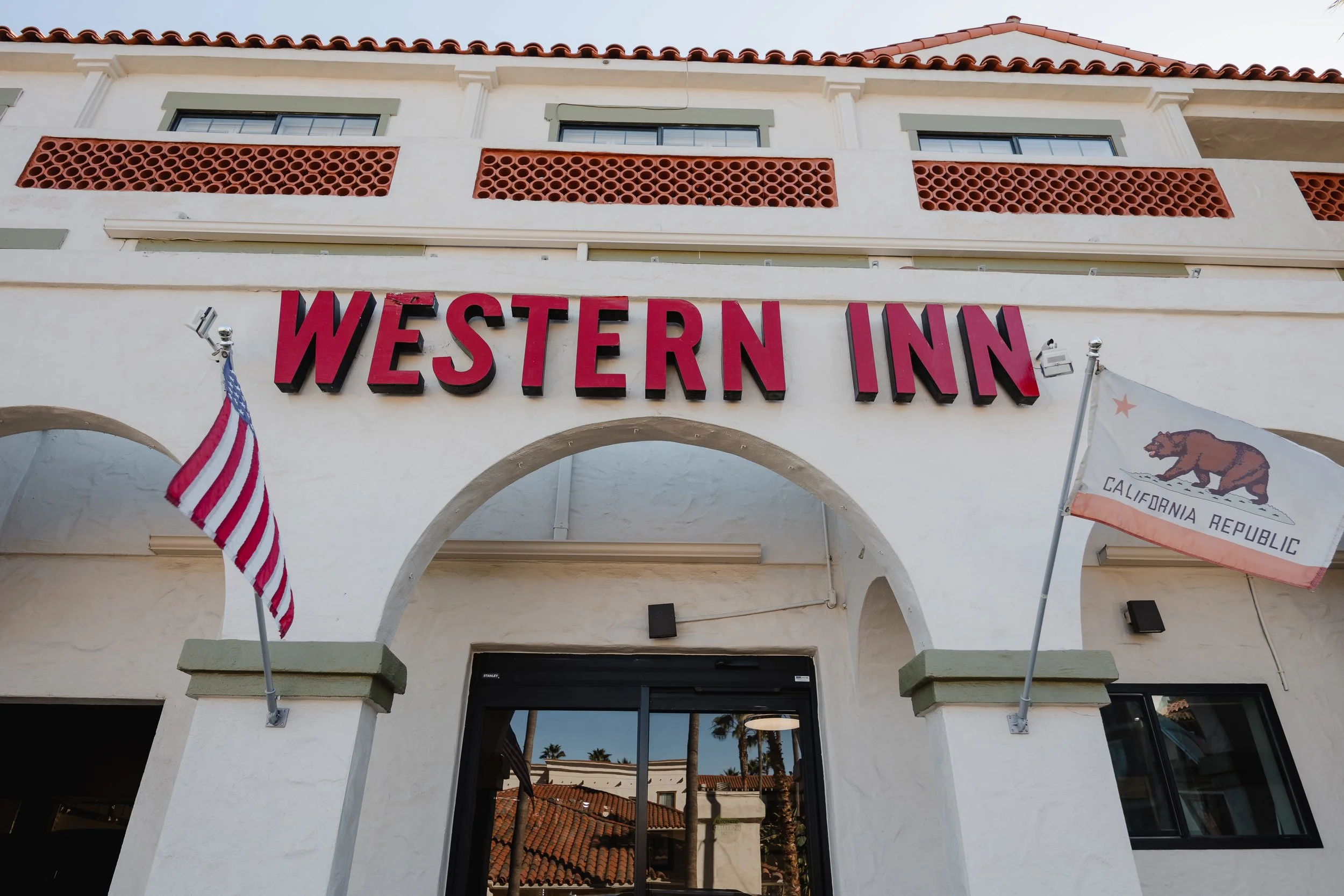 Exterior of a hotel building with a sign that reads 'Western Inn' and two flags, one American flag and one California state flag.