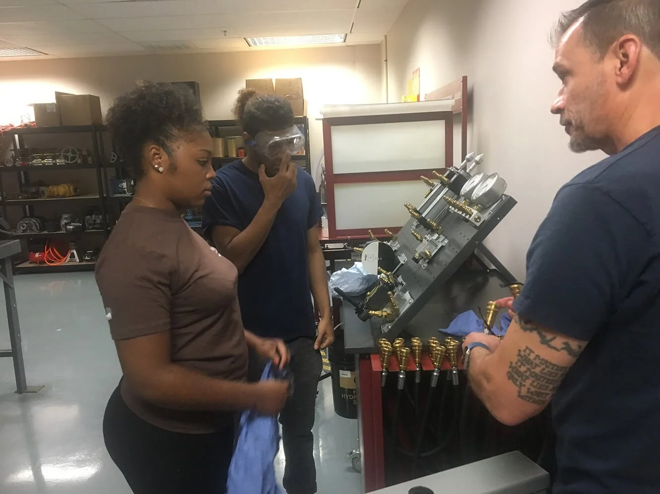 Three people observing a technician working on a panel with gauges and tubing in a workshop.