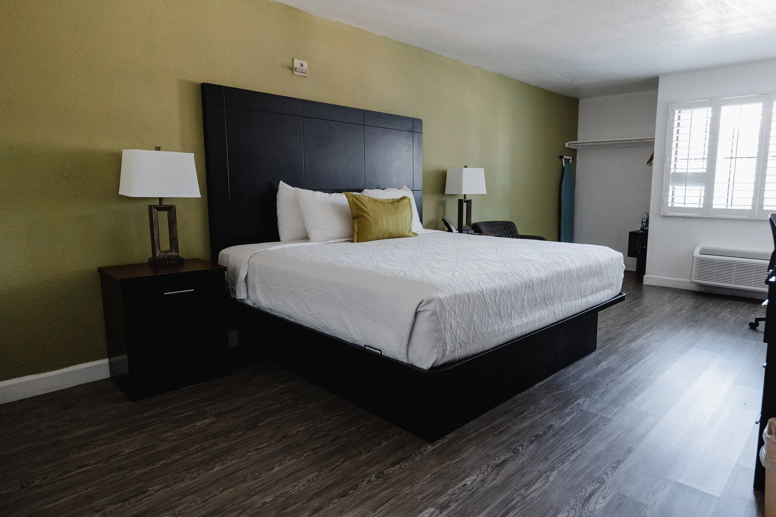 Hotel room with a large bed, nightstand with lamp, window with white shutters, and an ironing board in the corner.