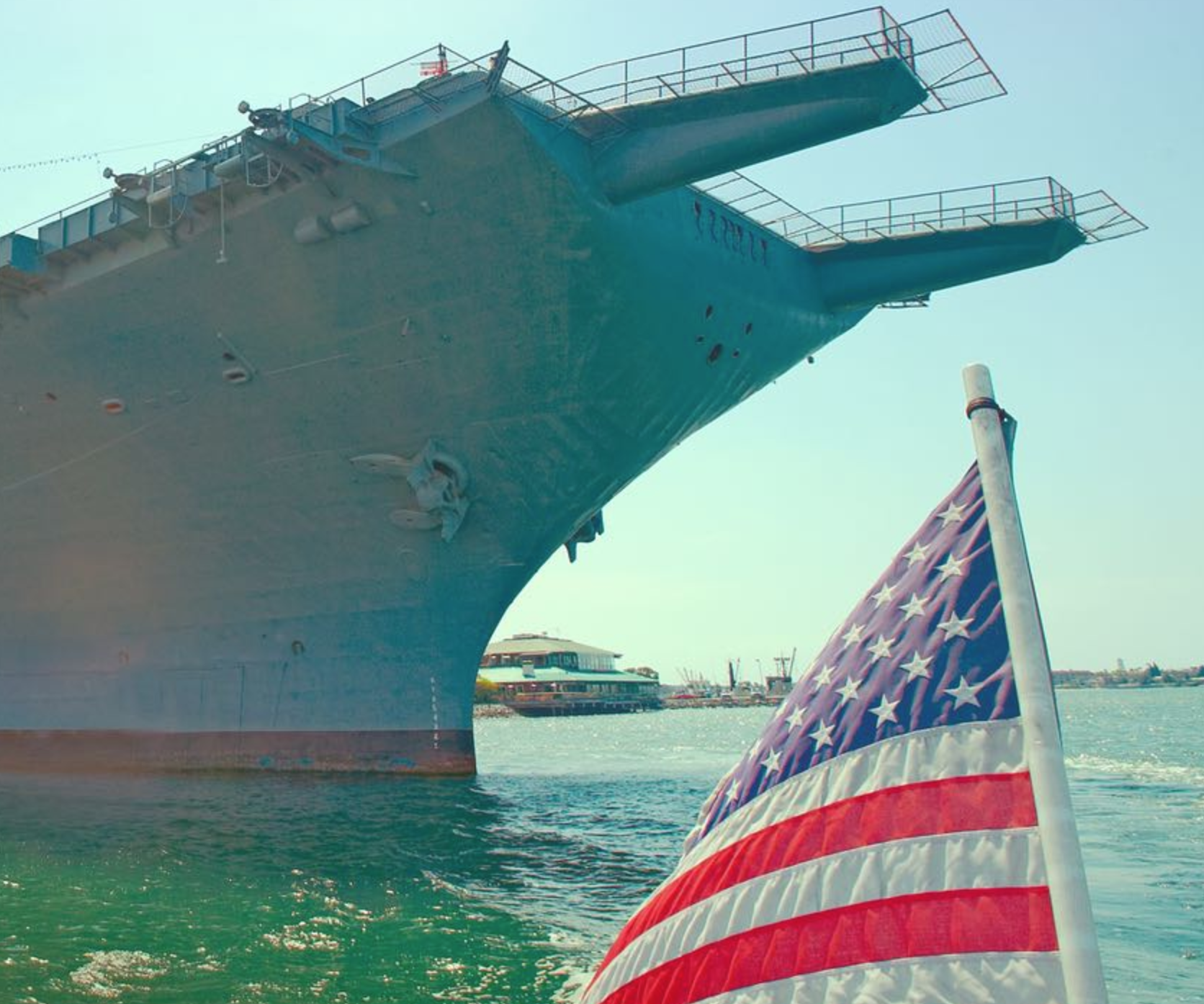 A large navy ship docked at a port with the bow visible, next to a waving American flag on a boat in the water, with other boats and port facilities in the background.