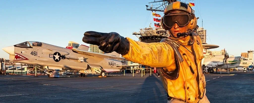 A military crew member in a yellow flight suit and helmet reaching out on an aircraft carrier with fighter jets in the background.