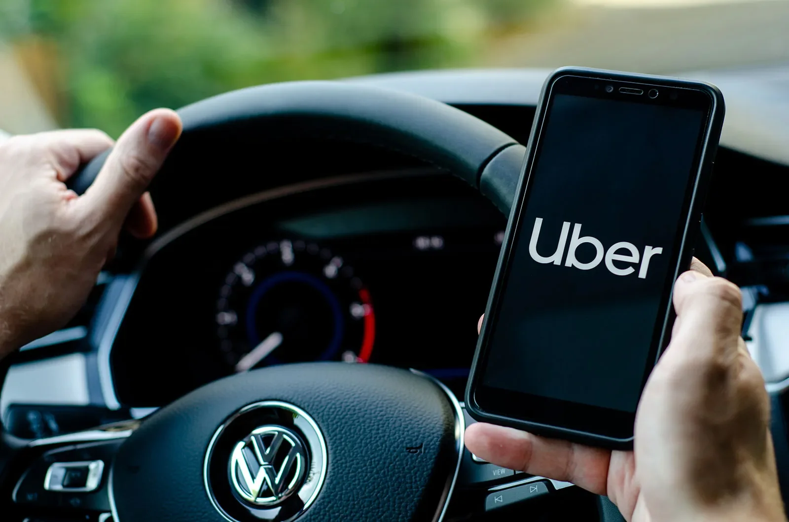 A person holding a smartphone displaying the Uber app while sitting in the driver's seat of a Volkswagen vehicle.
