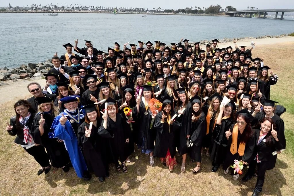 Group of graduates in caps and gowns celebrating outdoors near water, holding diplomas and flowers.