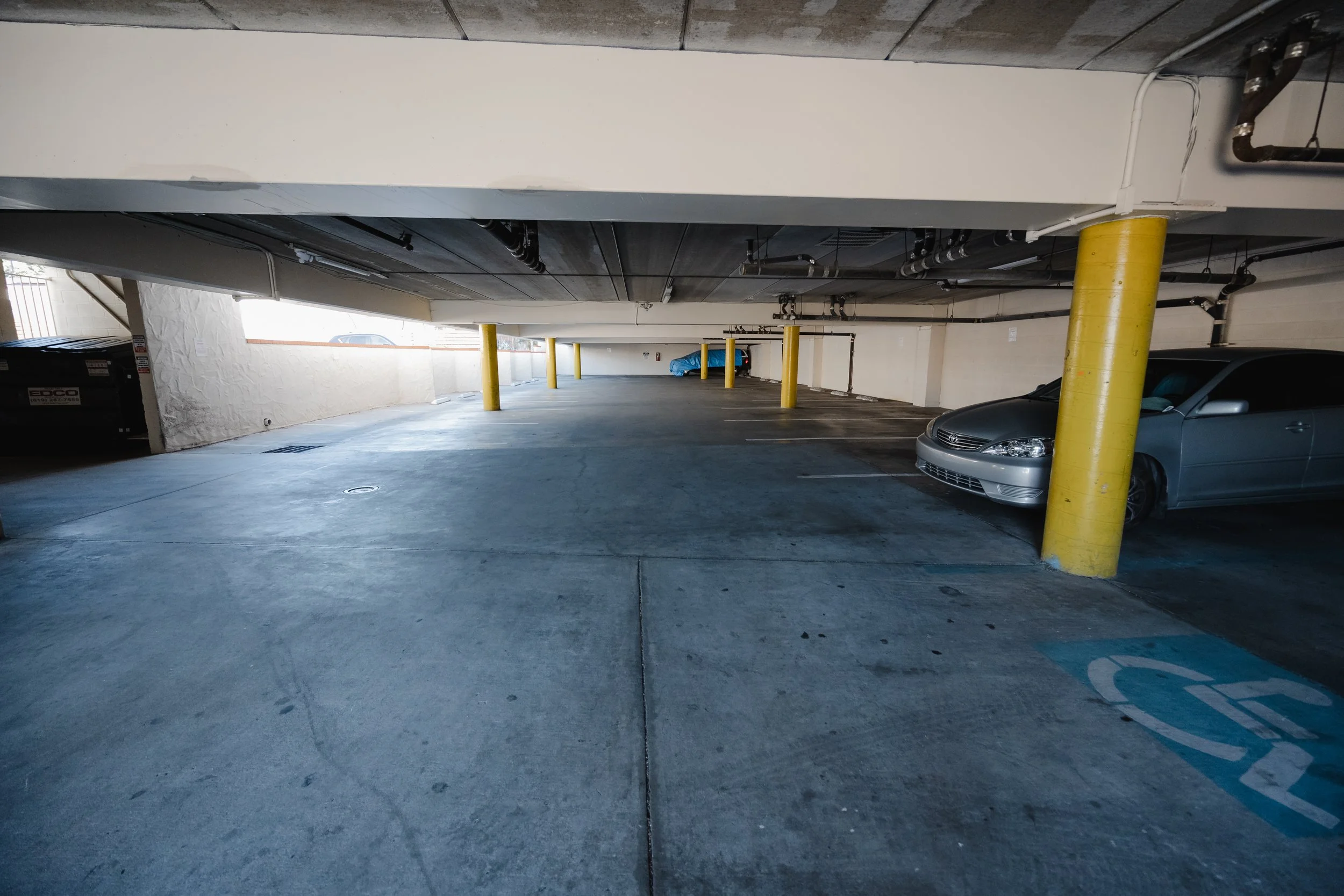Underground parking garage with yellow support columns, parked cars, exposed pipes, and a wash covered car in the background.