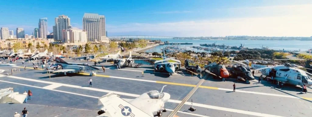 Various military aircraft parked on the deck of an aircraft carrier with a city skyline and a bay in the background.