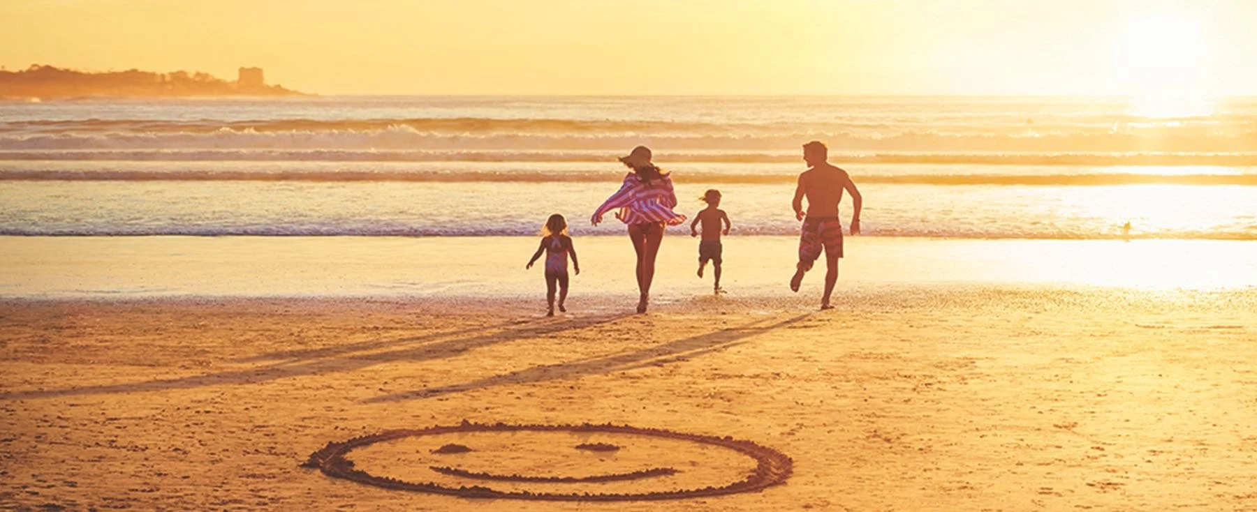 Family of four running on the beach at sunset, with a smiley face drawn in the sand.
