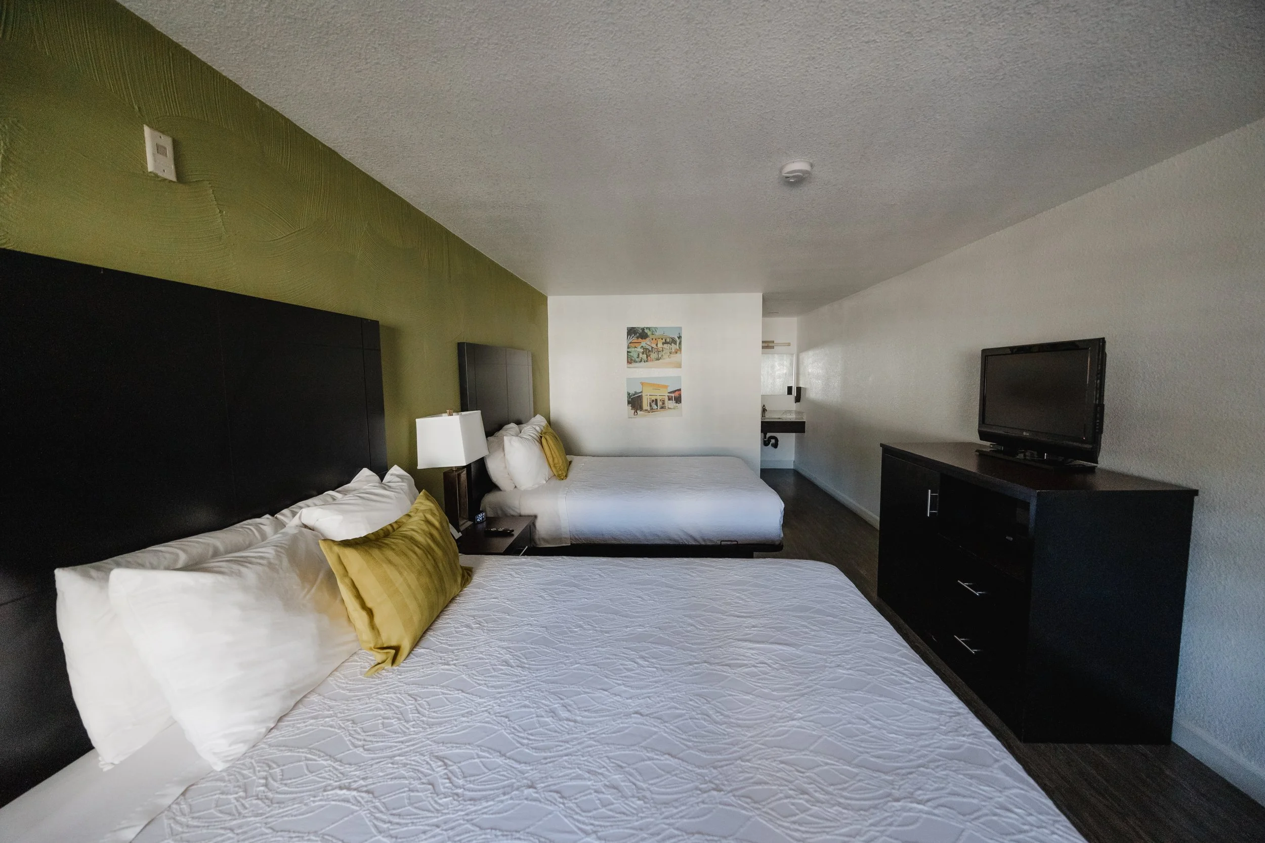 A hotel room with two beds, one with white bedding and yellow pillows, a black headboard, a white nightstand with a lamp, a flat-screen TV on a black dresser, and artwork on the wall.