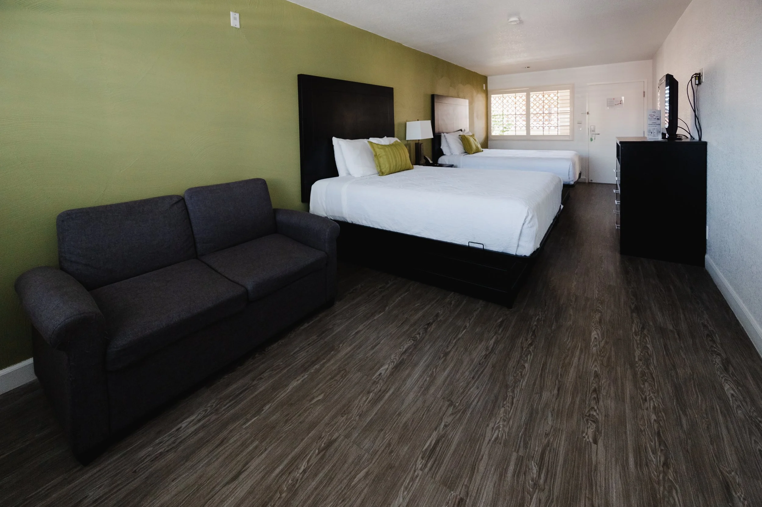 A hotel room with two beds against a green wall, a gray sofa, a window with blinds, and a dresser with a TV.