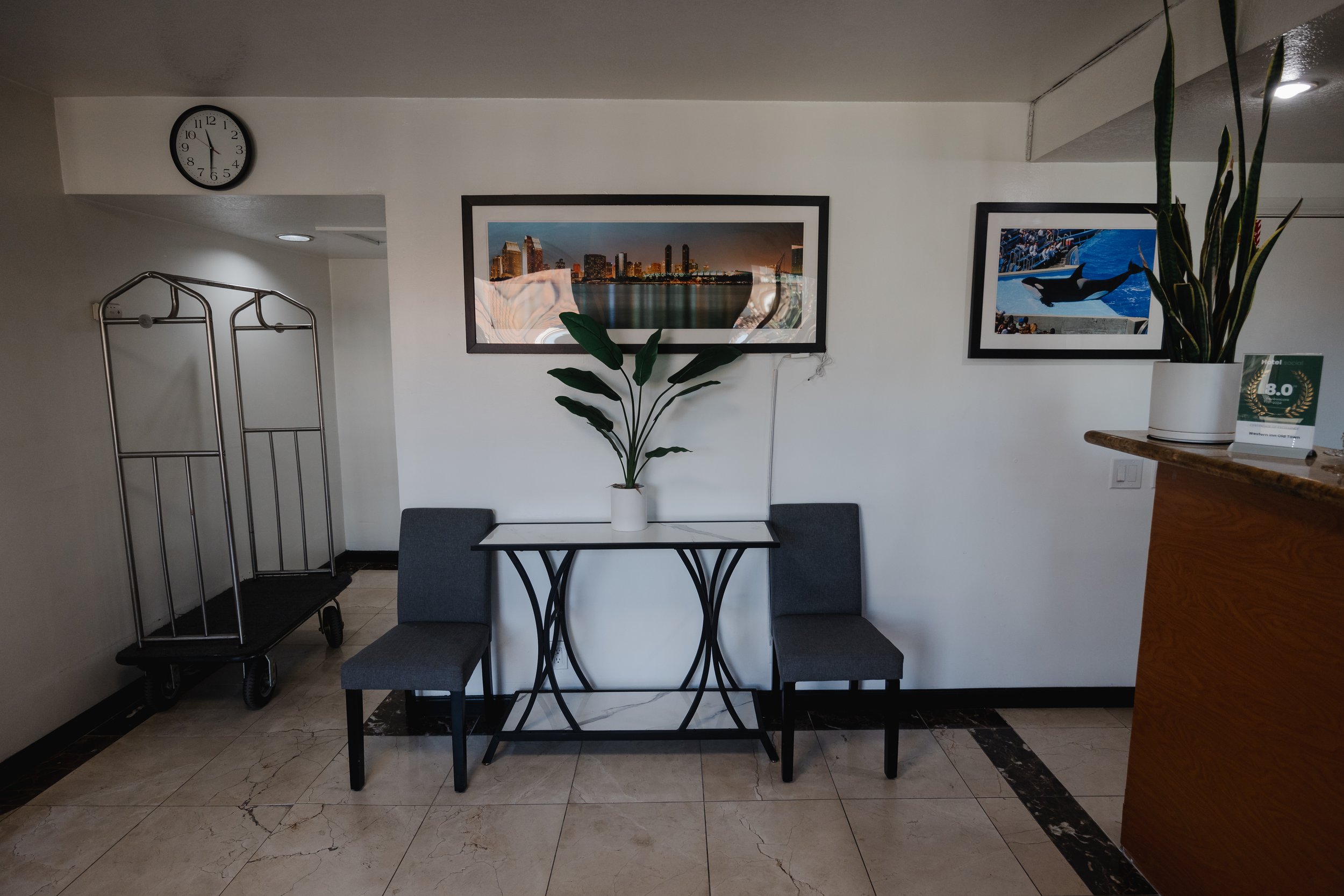 Interior of a room with a white wall, decorated with two framed cityscape and ocean photographs, a large potted plant, a small table, two chairs, a luggage cart, and a wall clock showing 10:55.