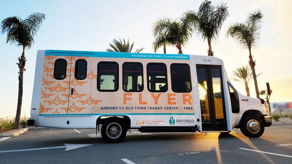 A bus labeled 'Flyer' in a parking lot with palm trees and a sunset sky in the background.