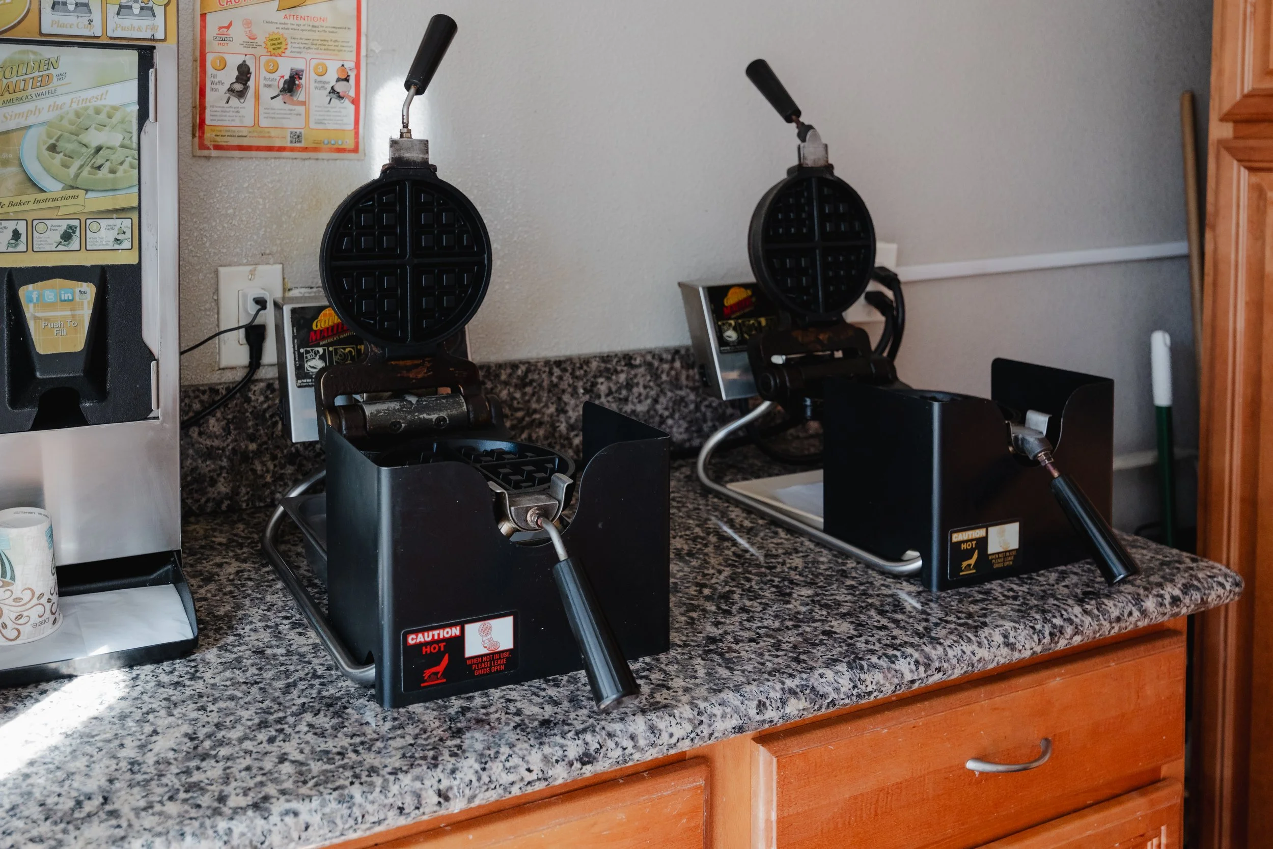 Two waffle makers on a granite countertop, one with a waffle in it and the other empty, with a scene from a restaurant or cafe kitchen.
