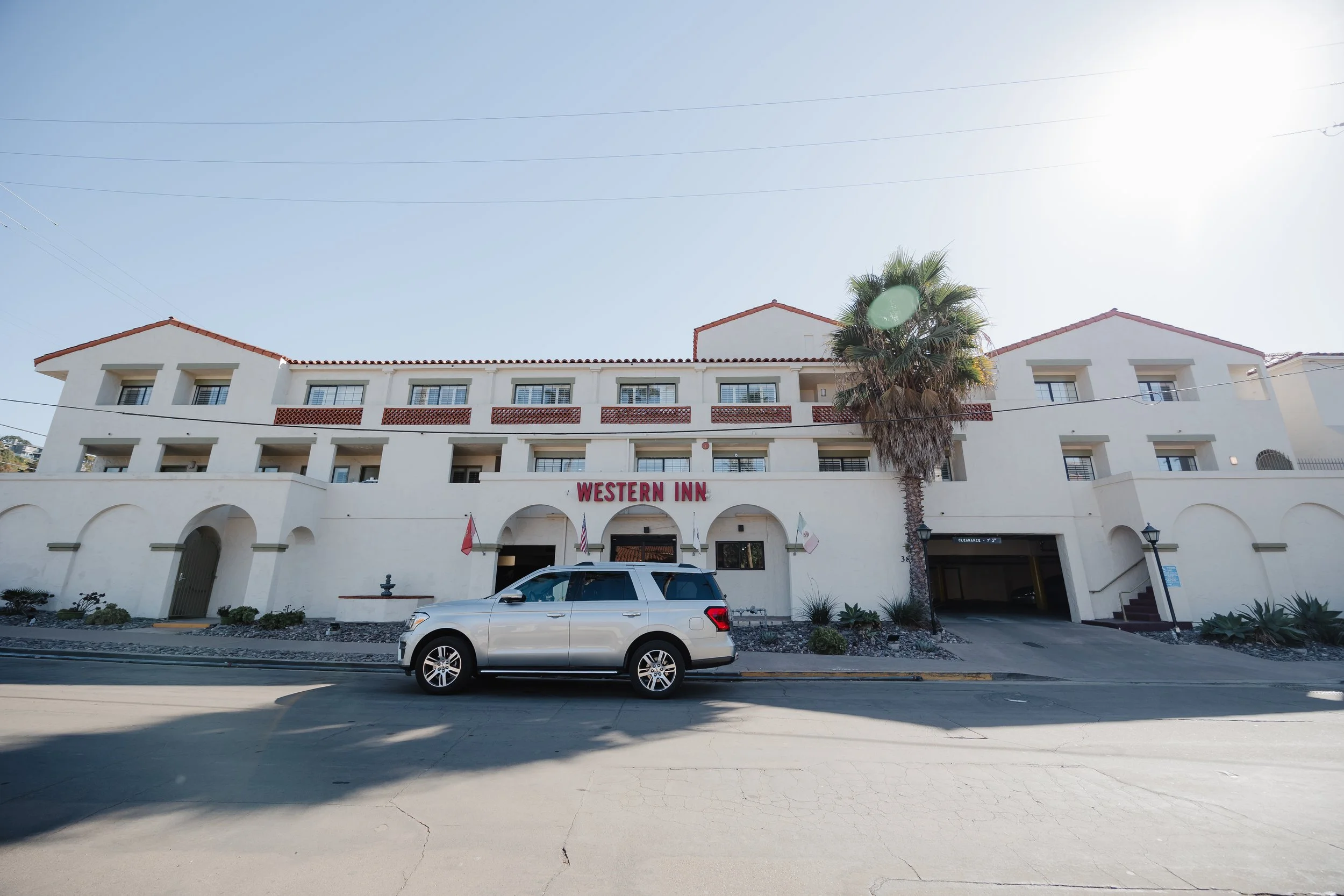 Front view of Western Inn hotel with a white building, red roof tiles, and a large palm tree, a silver SUV parked in front, and clear sunny sky.