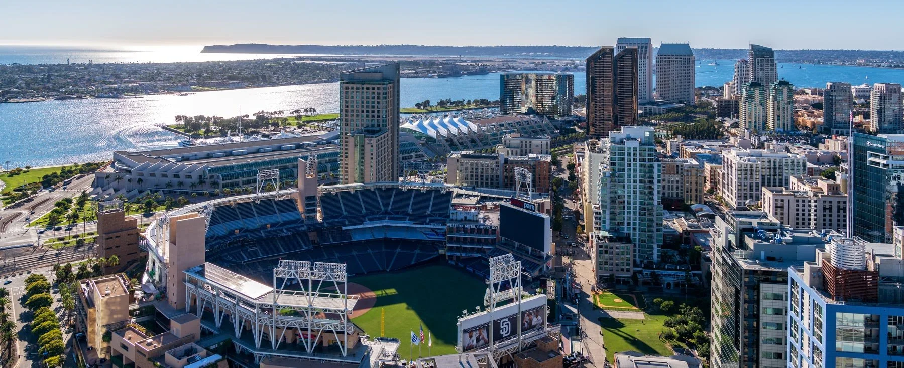 Aerial view of a city skyline with a stadium, high-rise buildings, and water in the background on a sunny day.