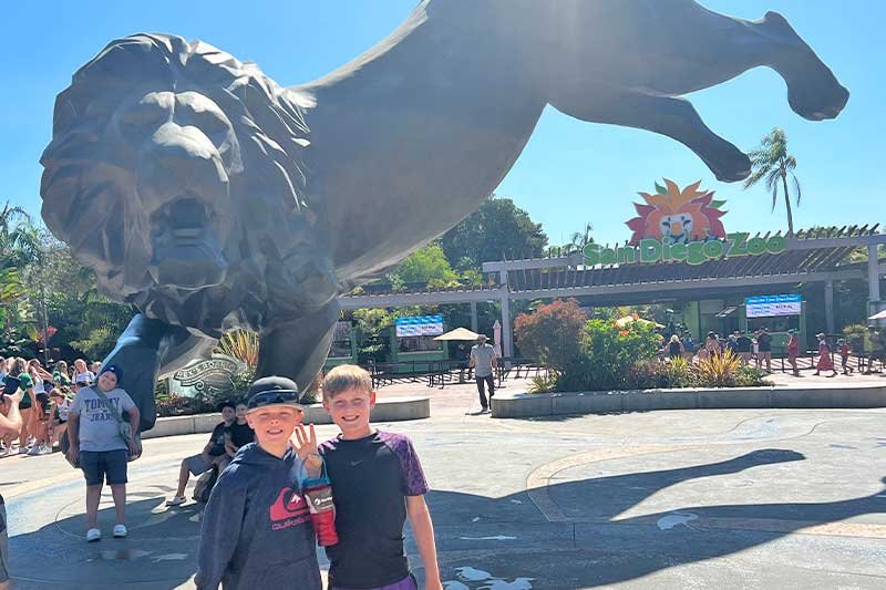Two boys standing in front of a large lion statue at the entrance of San Diego Zoo on a sunny day.
