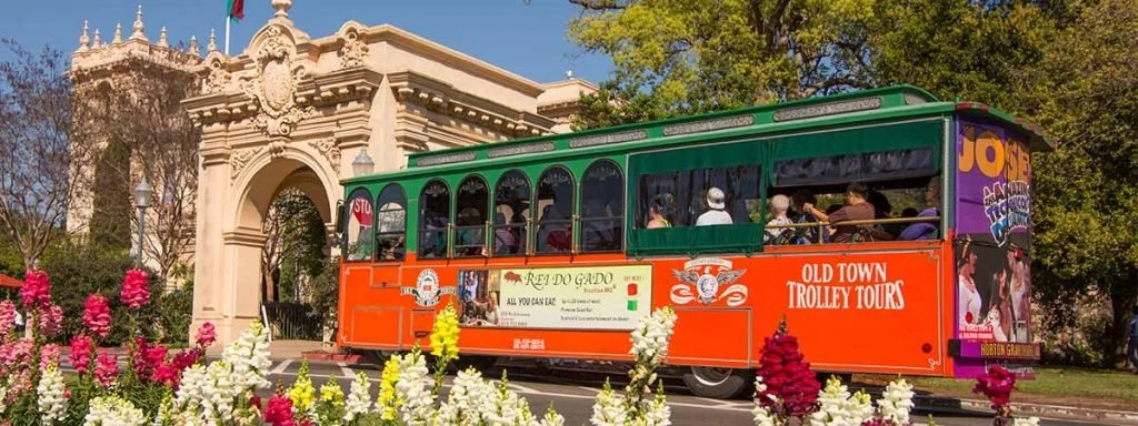 A colorful trolley with passengers, parked near a historic building and blooming flowers.