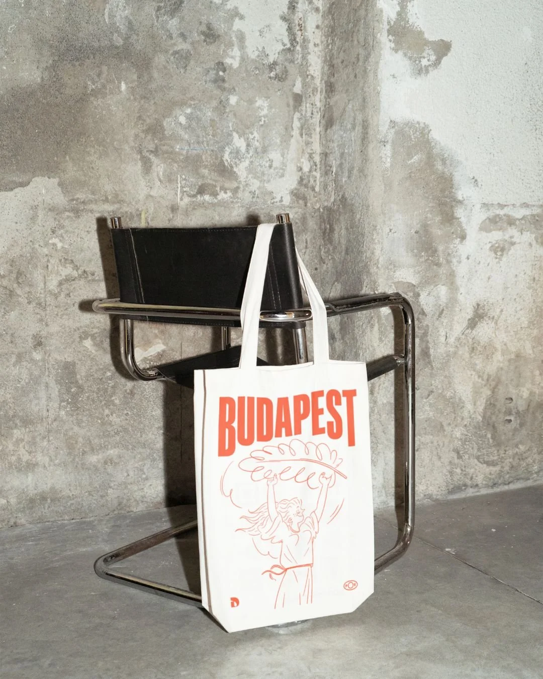 A white tote bag hanging on the armrest of a black and chrome chair, with the word 'BUDAPEST' in bold orange letters and a sketch of a woman holding a large leaf on the bag, set against a textured gray concrete wall.