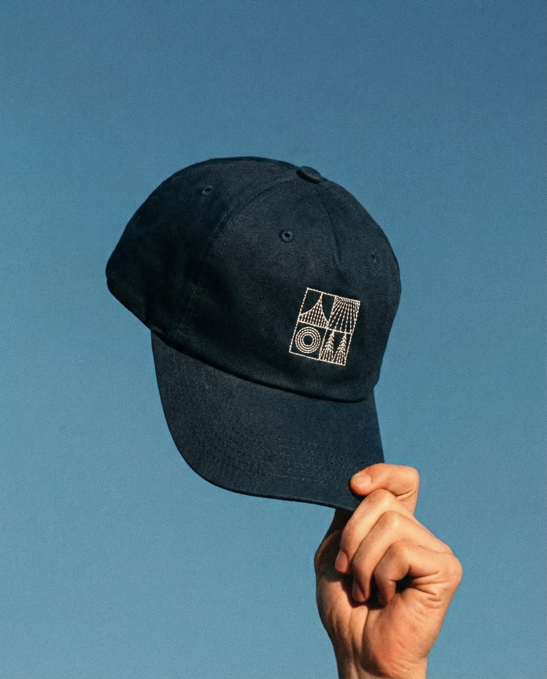 A person holding a navy blue baseball cap with a geometric design on the front, against a clear blue sky.