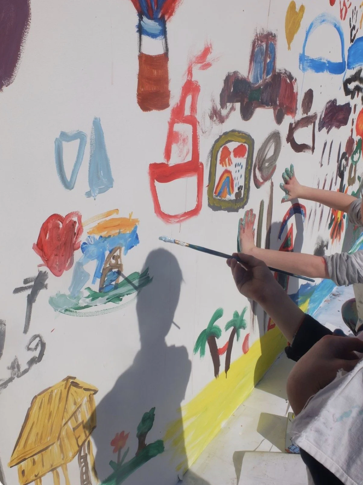Children painting and drawing on a large white wall with colorful paints and brushes.