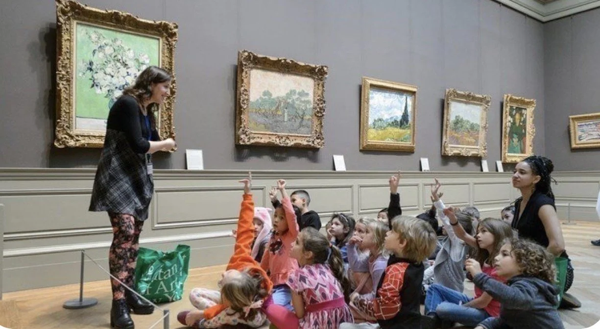 A group of children sitting on the floor in an art museum, listening and raising their hands, while a museum guide or teacher stands in front of them with artworks displayed on the wall.
