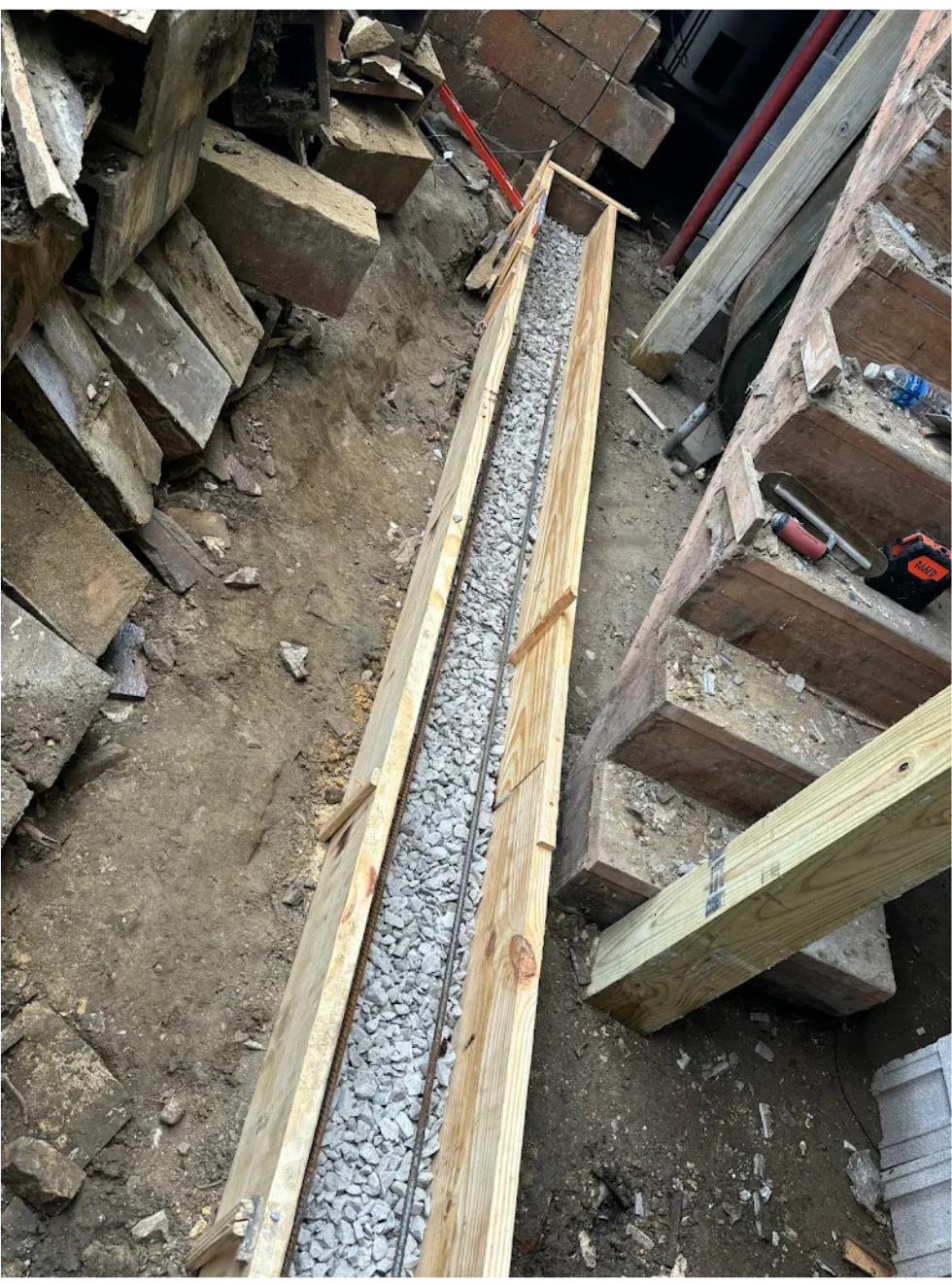 Construction site showing a narrow trench with gravel and rebar, framed with wooden boards for concrete work, surrounded by construction tools and materials.