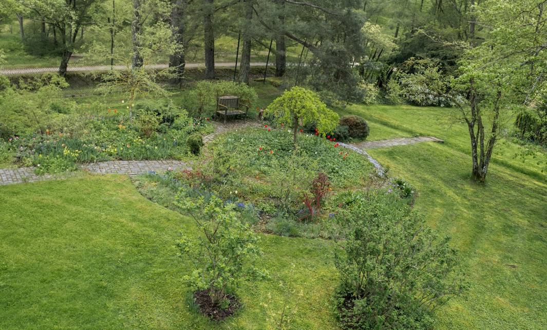A lush, green backyard garden with flowering plants, trees, a bench, and a winding stone pathway.