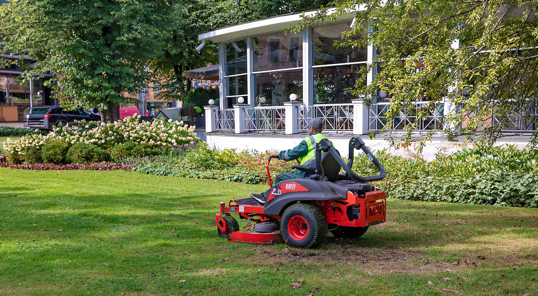 A person riding a red and black ride-on mower on a grassy lawn in front of a building with large windows and a porch, surrounded by trees and shrubs.