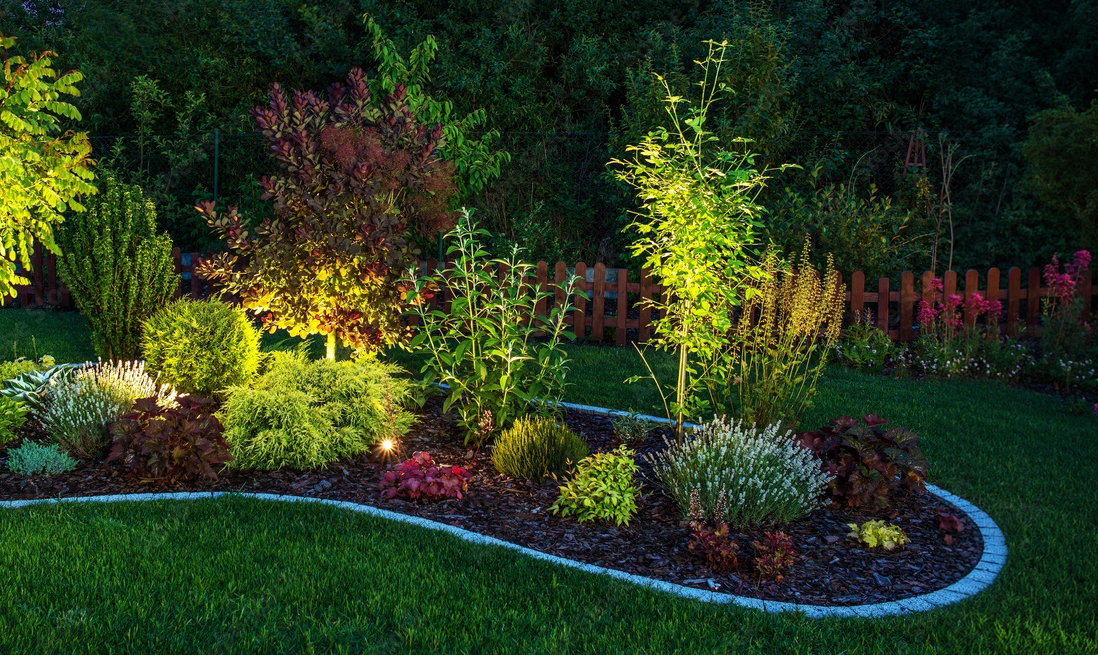 Nighttime garden scene with illuminated various plants, shrubs, and flowers in a landscaped flower bed surrounded by well-maintained grass and a wooden fence in the background.