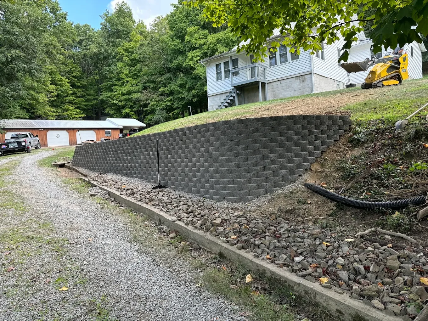A newly constructed retaining wall made of interlocking concrete blocks on a sloped yard, with a gravel pathway beside it and a house with a white exterior and garage in the background.