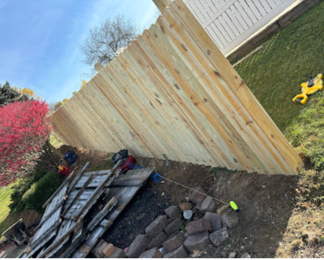 A newly built wooden fence in a backyard with construction tools and materials around, including a wheelbarrow, bricks, and a small yellow toy. There are trees and bushes nearby, with a blue sky overhead.