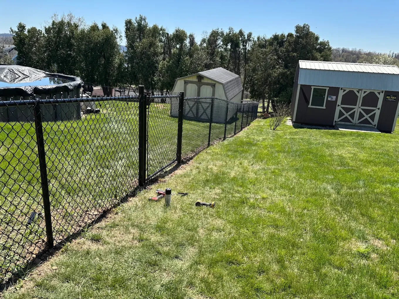 Yard with a black chain-link fence, a shed, a storage barn, and a trampoline, with trees in the background and a blue sky.