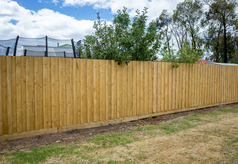 New wooden privacy fence in a backyard with trees and a trampoline in the background, under a partly cloudy sky.