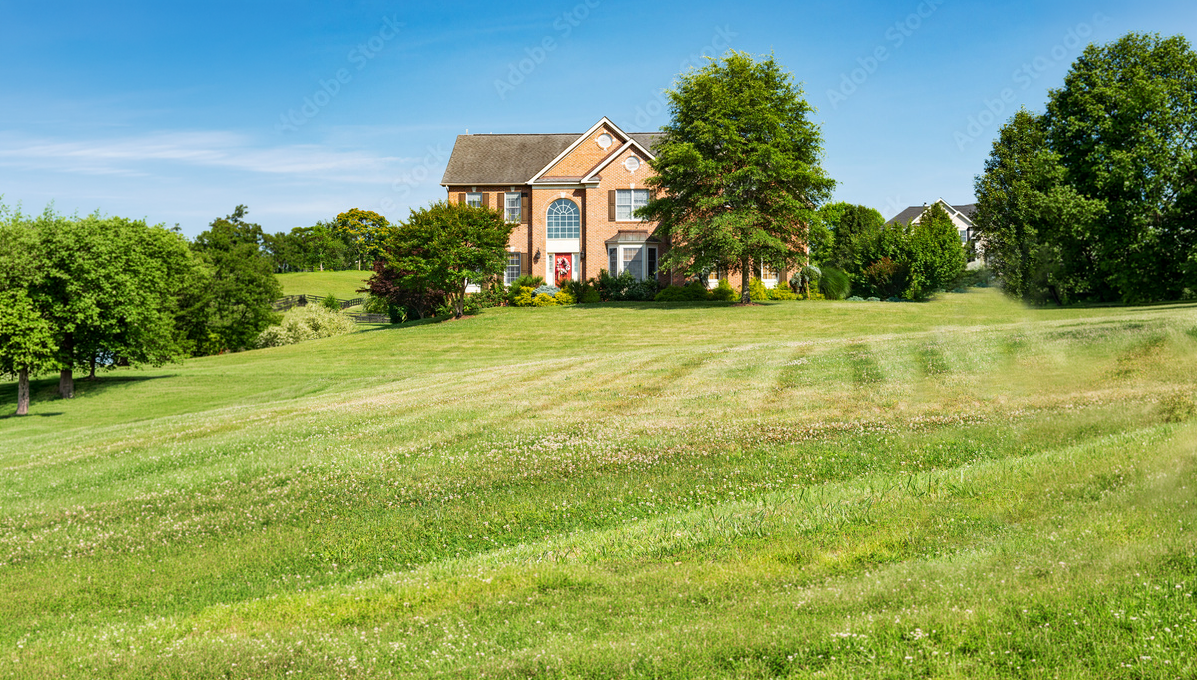 A large brick house surrounded by green trees and a well-maintained lawn under a bright blue sky.