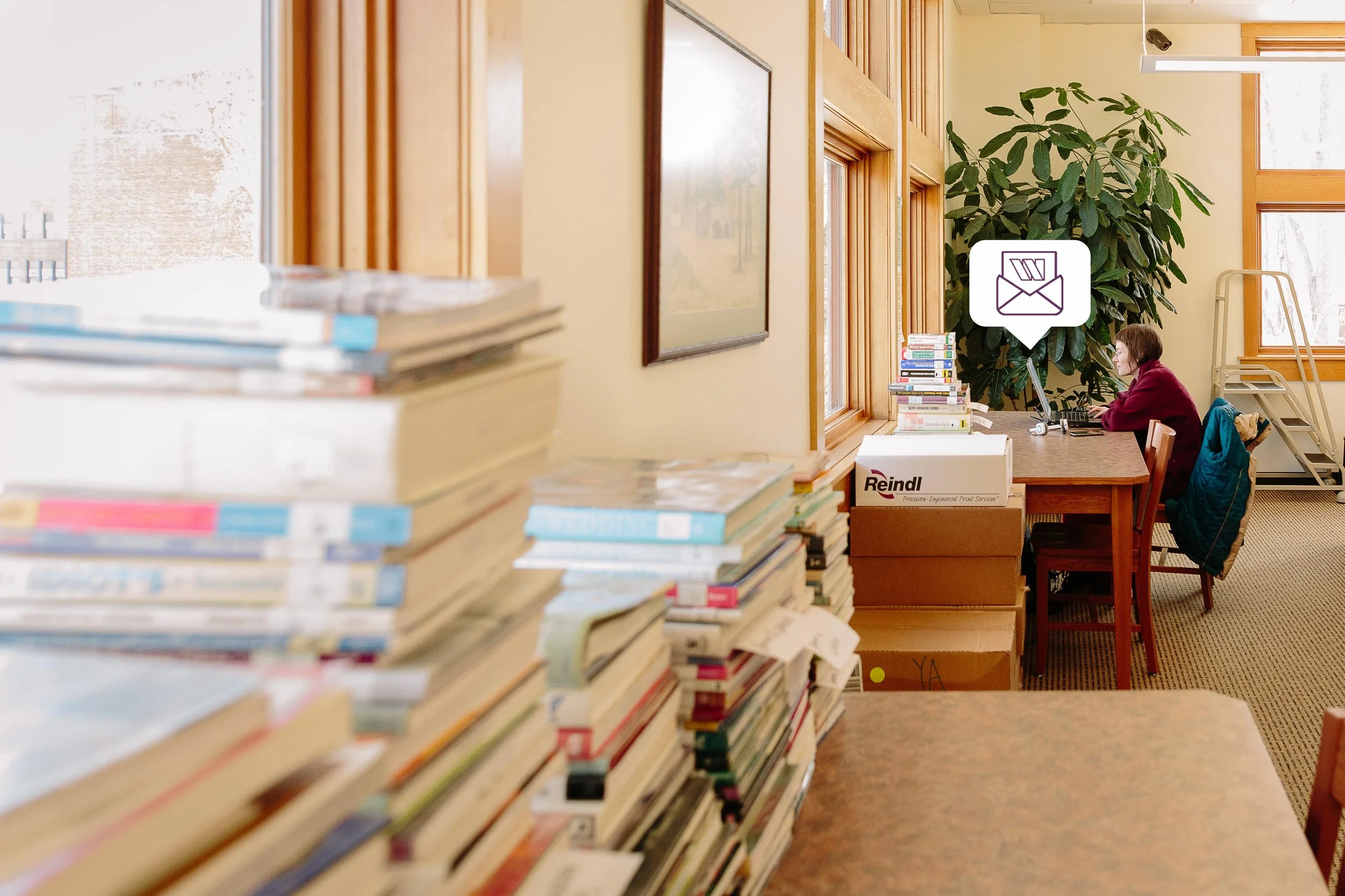 A woman in the distance working on a laptop with a tall stack of books in front of her. There is also a few stacks of books in the foreground on a closer table.