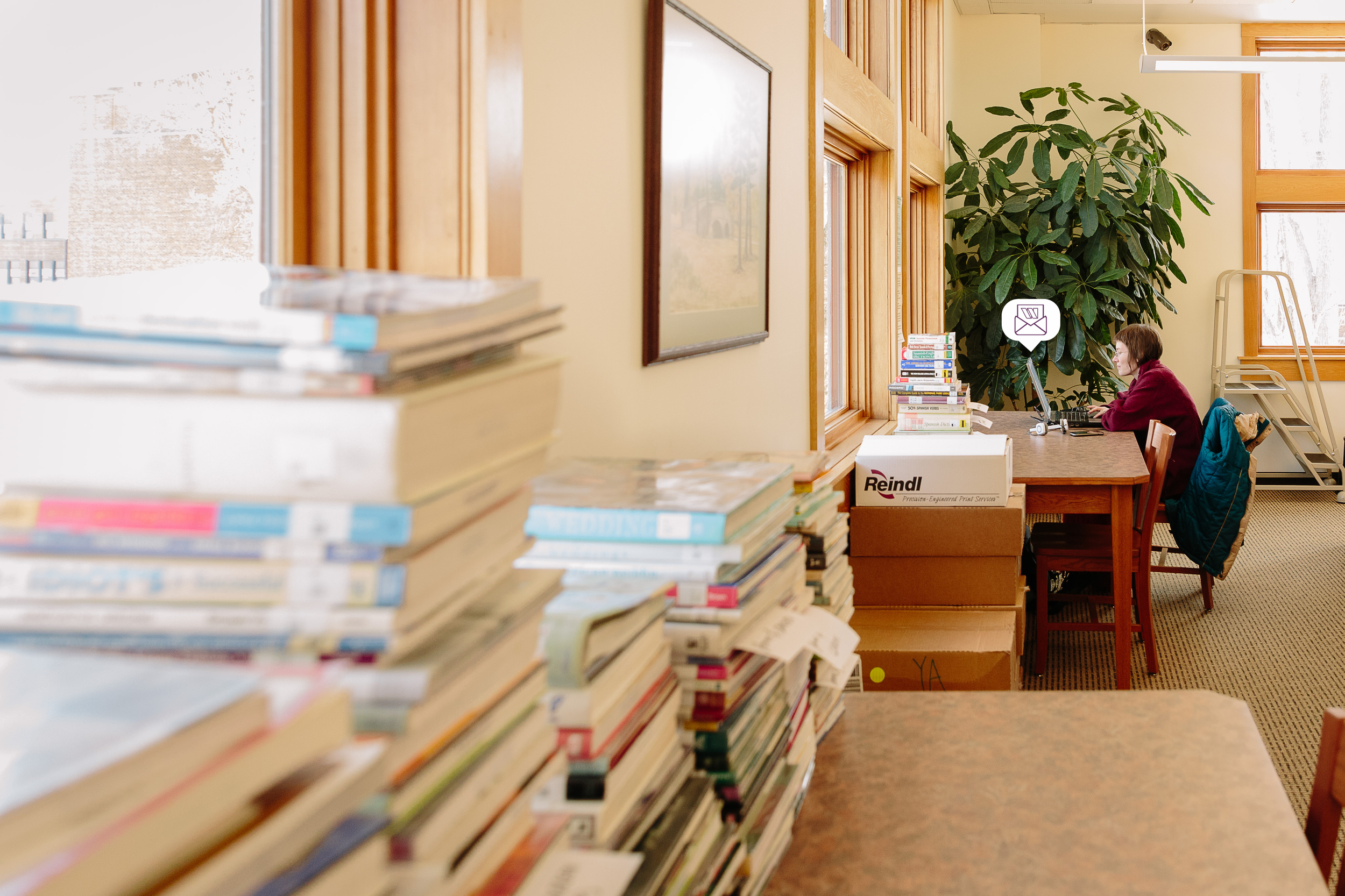 A woman in the distance working on a laptop with a tall stack of books in front of her. There is also a few stacks of books in the foreground on a closer table.