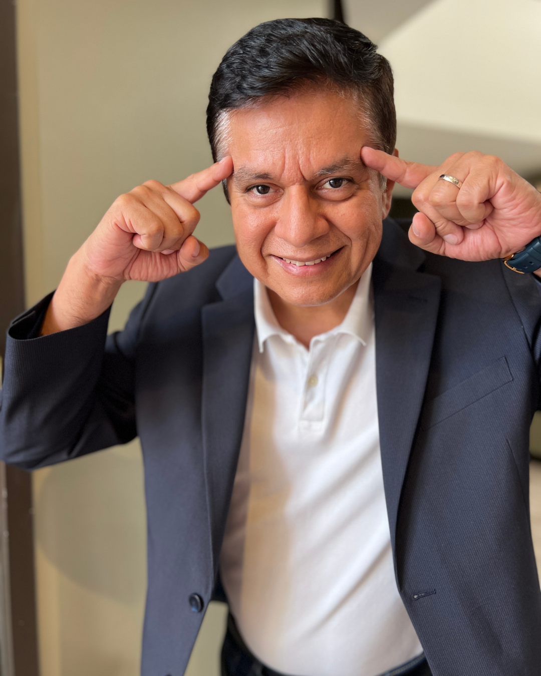 A smiling man in a suit pointing to his temples with both index fingers.
