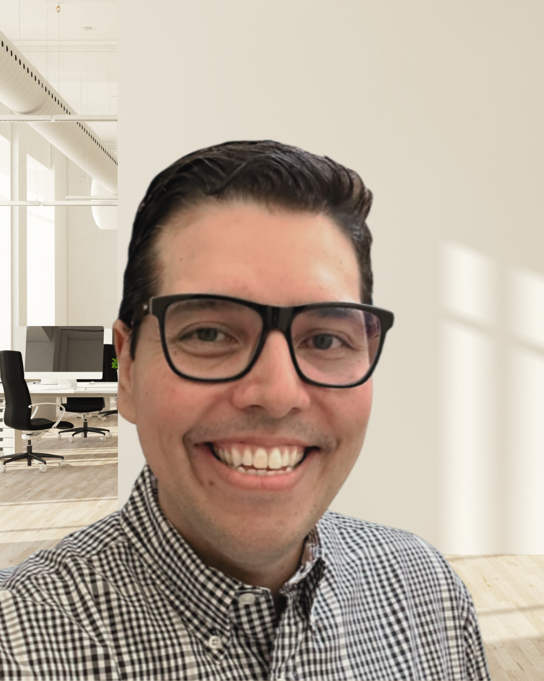 A smiling man with short dark hair, wearing glasses and a checkered shirt, taking a selfie in a modern office with white walls, large windows, and a workspace with chairs and computers in the background.