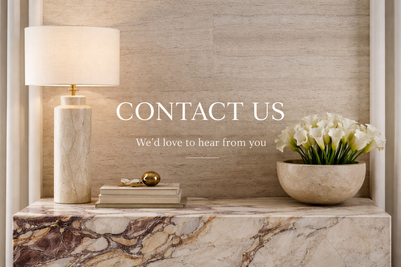 Interior decor with a table lamp, a stack of books, a decorative gold sphere, and a bowl of white calla lilies, with the text "Contact Us. We'd love to hear from you."