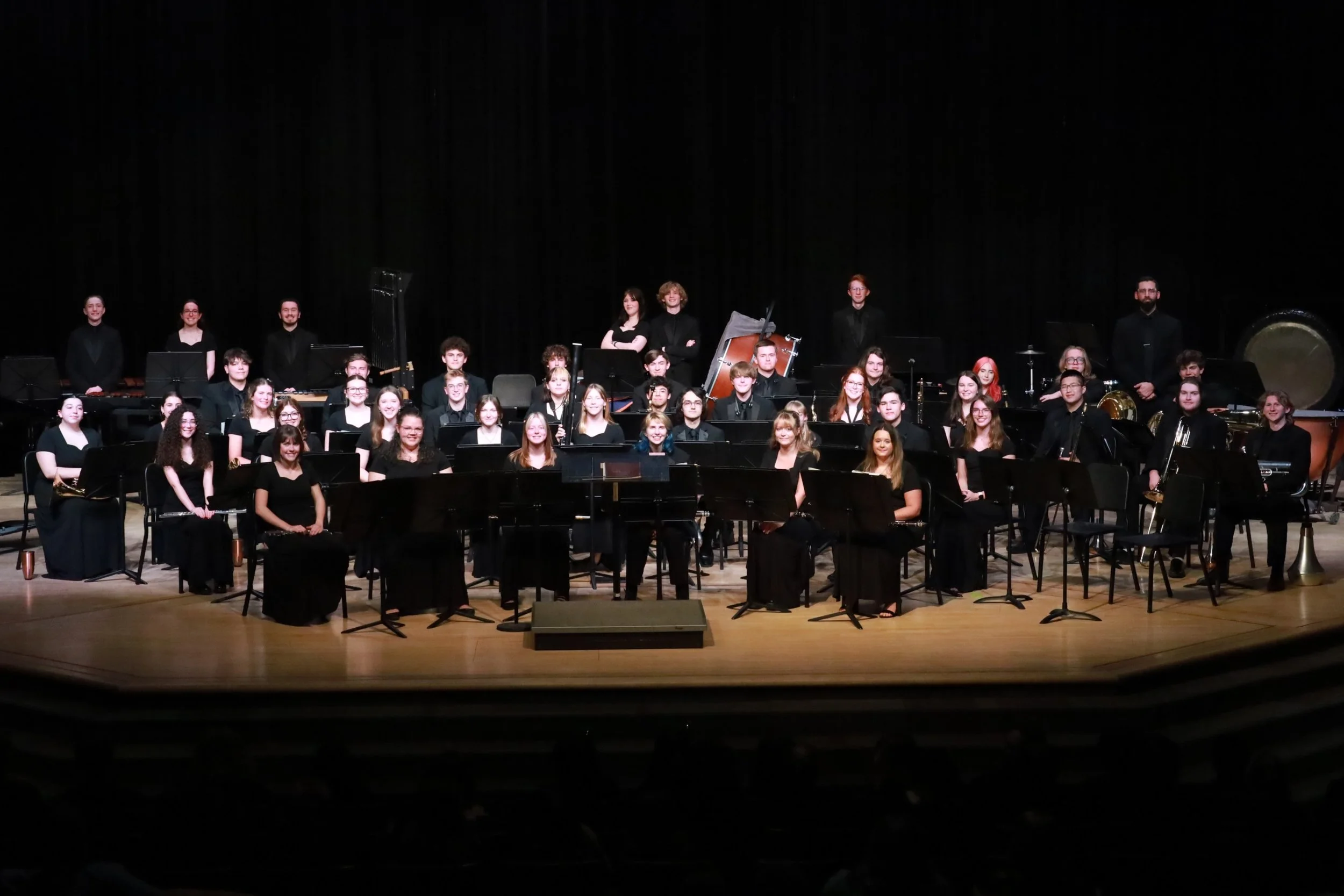 Band of young musicians on stage with black backdrop, seated with their musical instruments, ready for performance.