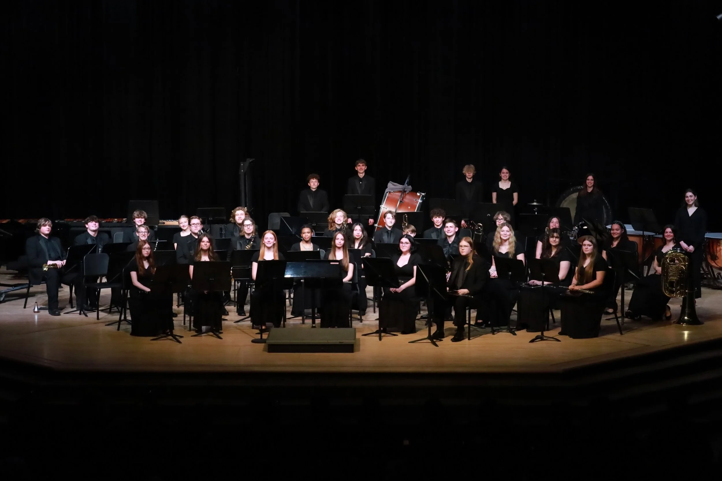 Group of young musicians in formal black attire sitting and standing with musical instruments on stage in an auditorium.