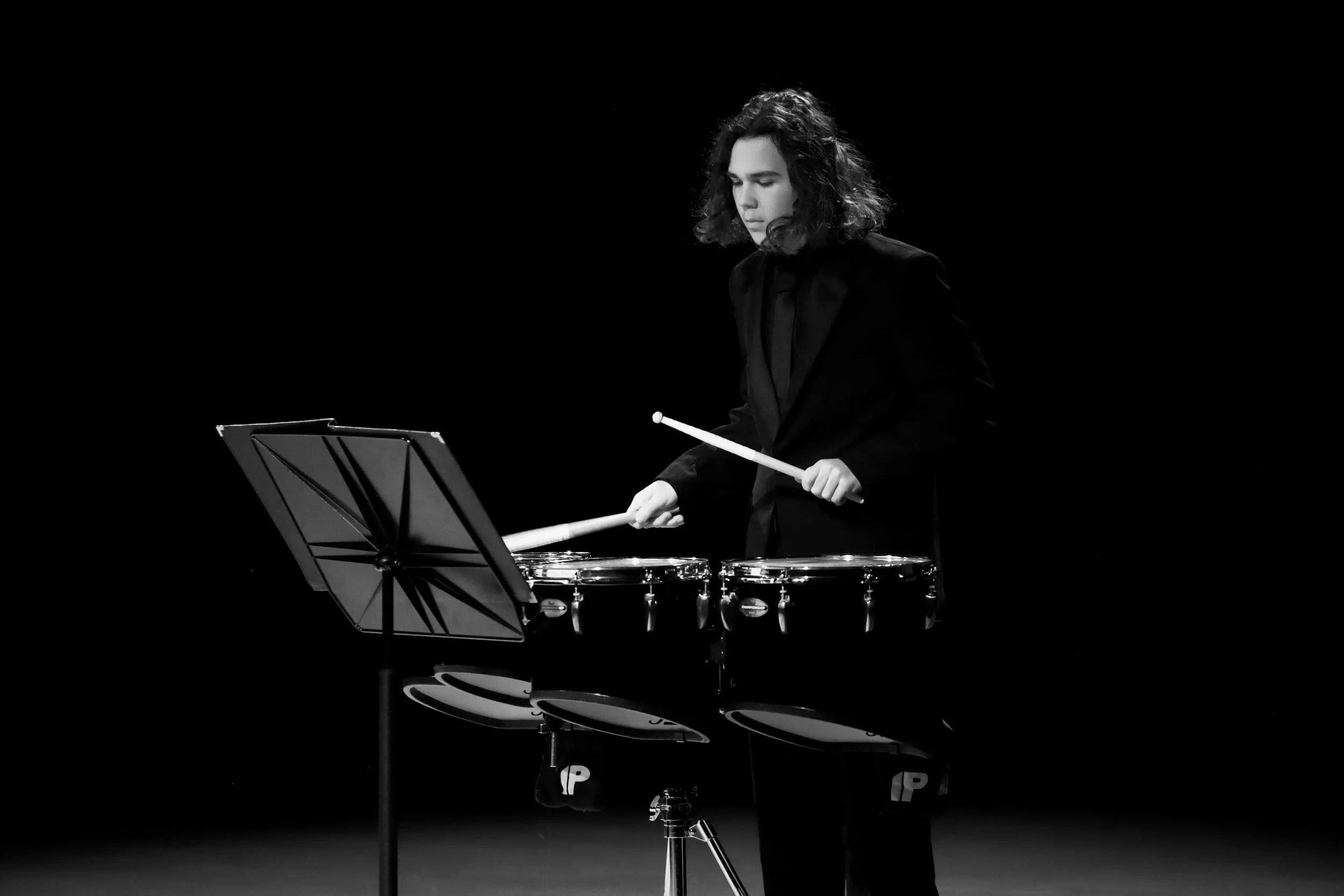 A young man with long hair wearing a black tuxedo plays quad drums with drumsticks on a dark stage.