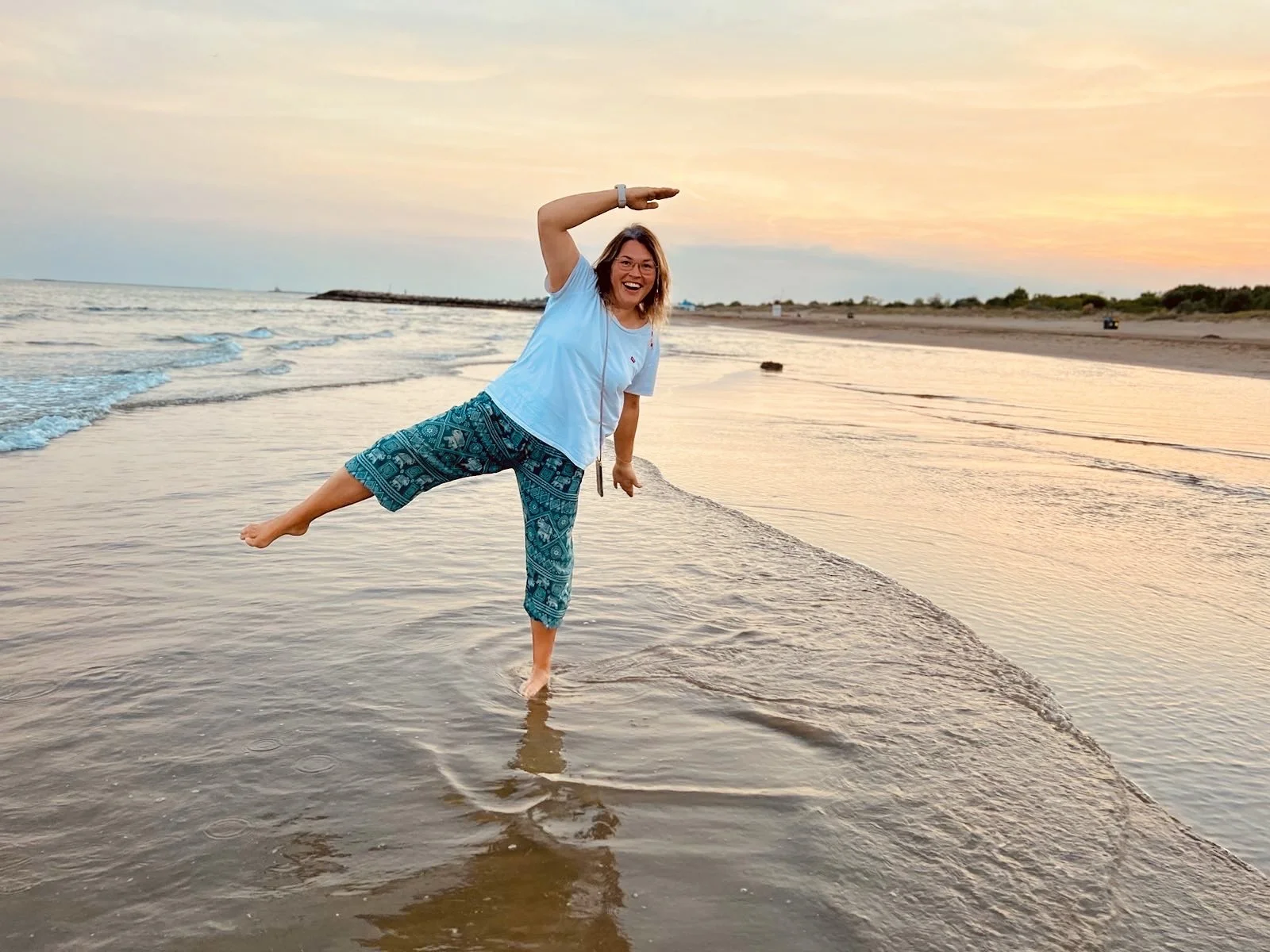 Eine lachende Frau in bunten Hosen und einem weißen T-Shirt steht im Wasser am Strand bei Sonnenuntergang, balanciert auf einem Bein und winkt mit der Hand über ihrem Kopf.