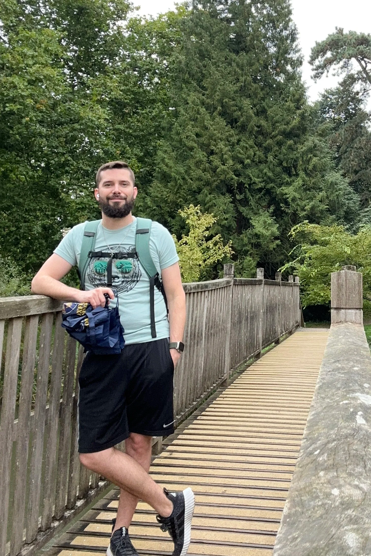 A man with a beard and short brown hair, wearing a light blue T-shirt with a face illustration, black shorts, black sneakers, a backpack, and earbuds, standing on a wooden bridge with green trees in the background.